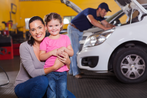 Smiling woman hugging smiling little girl while technician inspect white vehicle in background