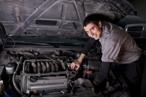 Caucasian mechanic in grey overalls and black long sleeve tee looking under the hood of a vehicle