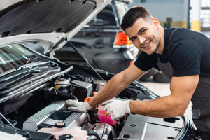 Smiling Mechanic wearing black tee and white gloves looking under a vehicle's hood